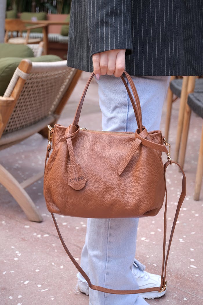 A stylish brown leather handbag held by a person in a casual outdoor setting.