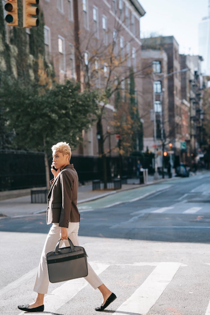 Full body side view of African American female entrepreneur in casual outfit with laptop bag having phone call while crossing road